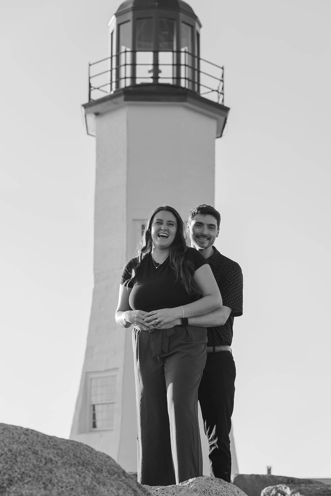 Black-and-white portrait of an engaged couple smiling together beneath Scituate Lighthouse