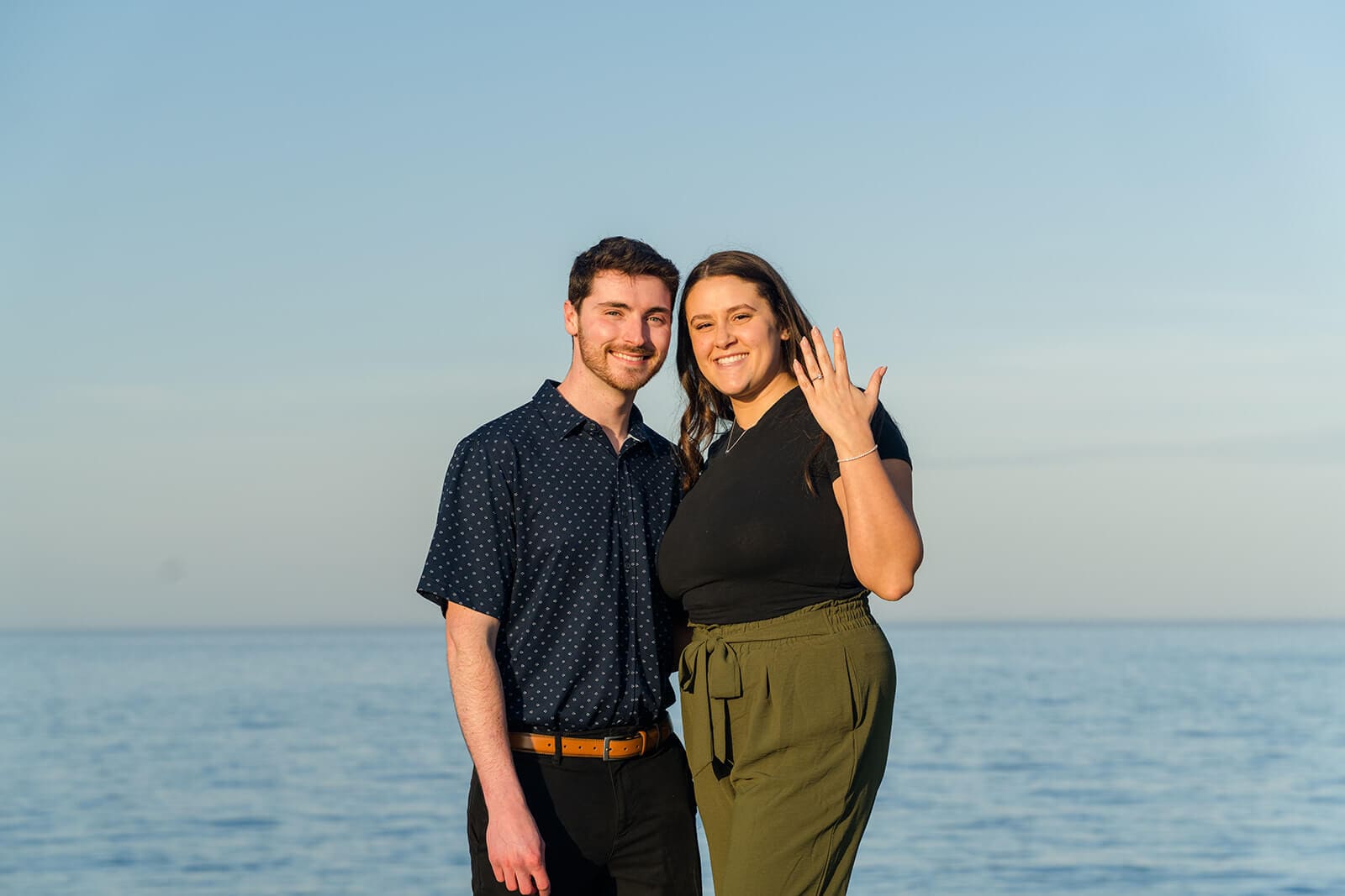 Newly engaged couple smiling together with the ocean behind them as the woman shows her ring