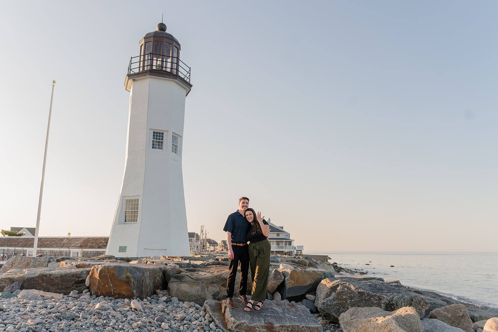 Engaged couple standing on coastal rocks with Scituate Lighthouse towering behind them