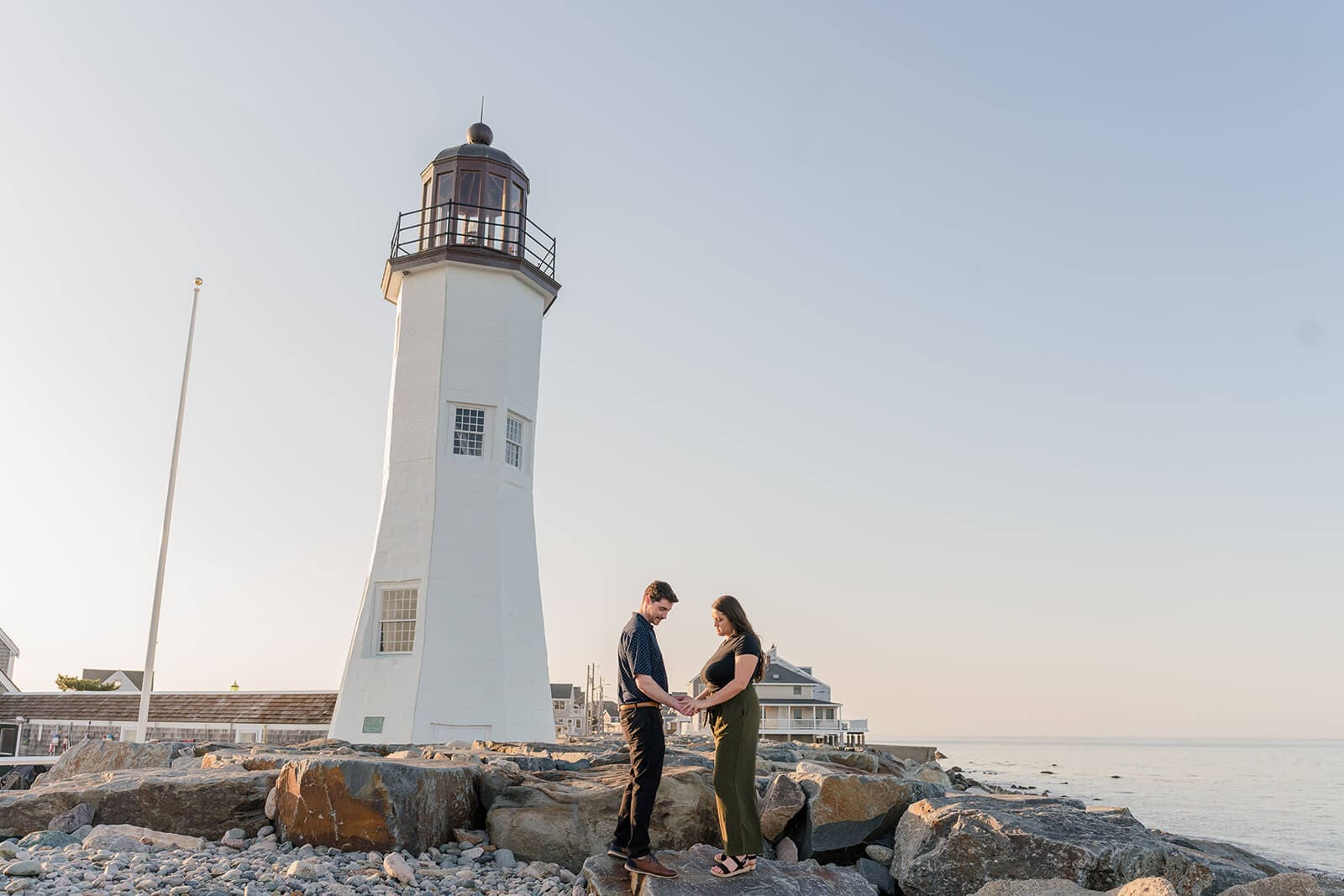 Couple looking down at the engagement ring together on the rocks in front of Scituate Lighthouse