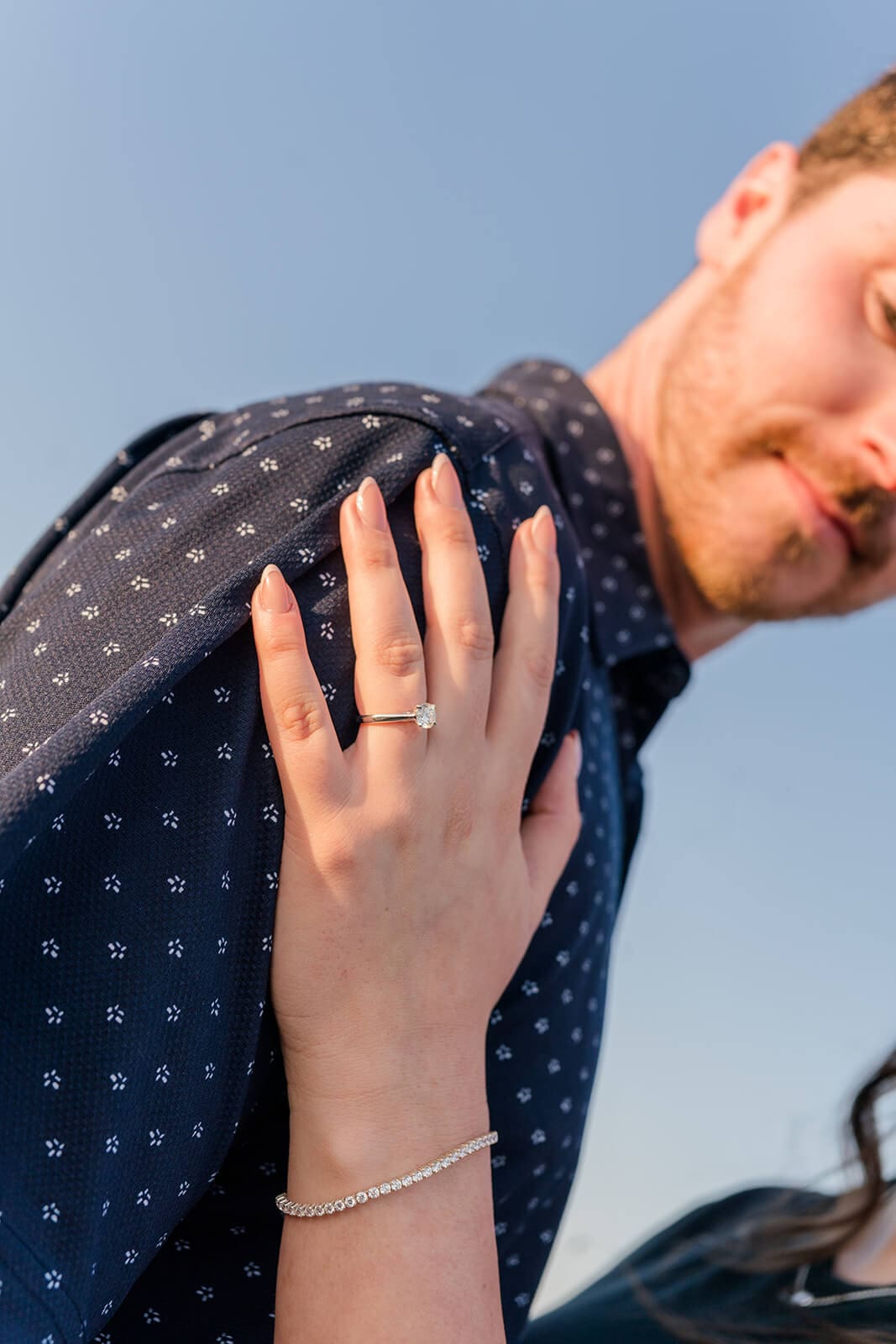 Close-up of a woman’s engagement ring as her hand rests on her fiancé’s shoulder