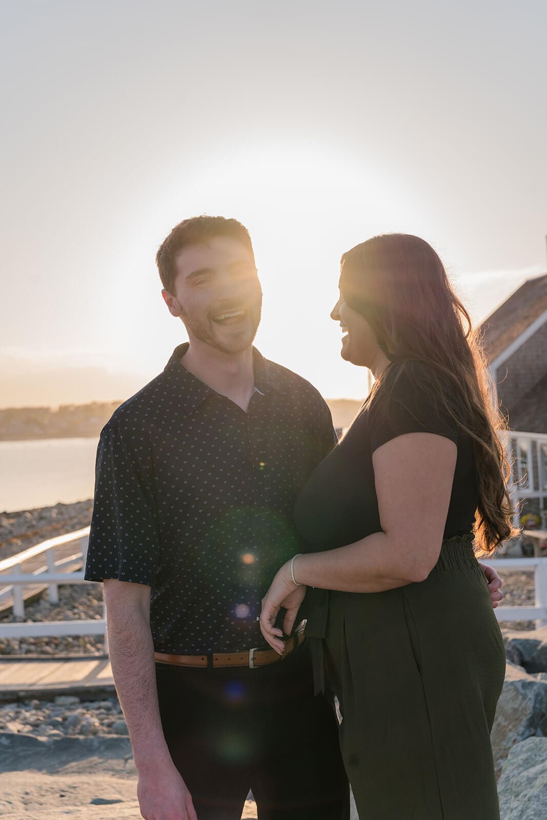 Engaged couple smiling at each other in warm sunset light near the water