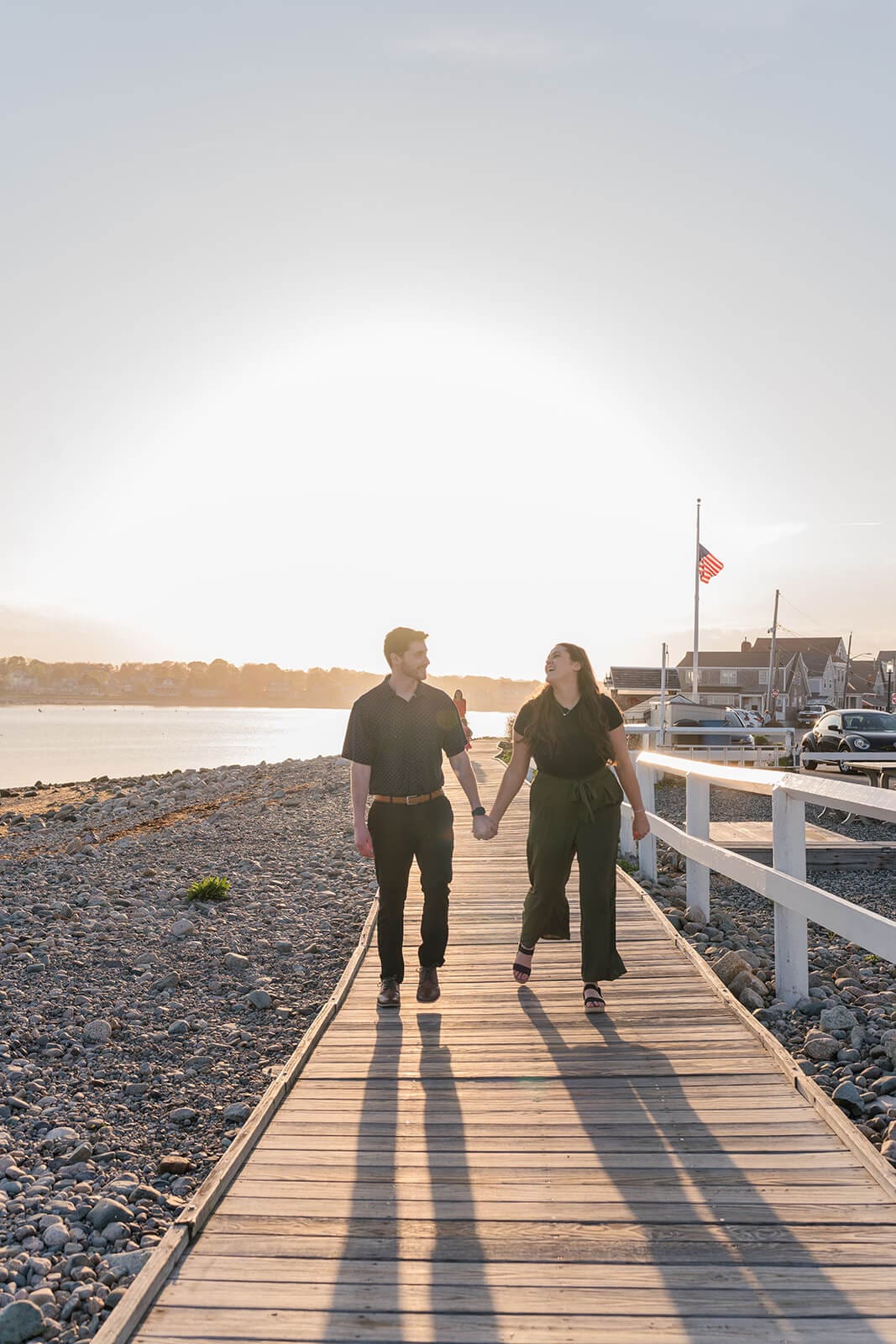 Newly engaged couple walking side by side along a waterfront boardwalk in golden evening light