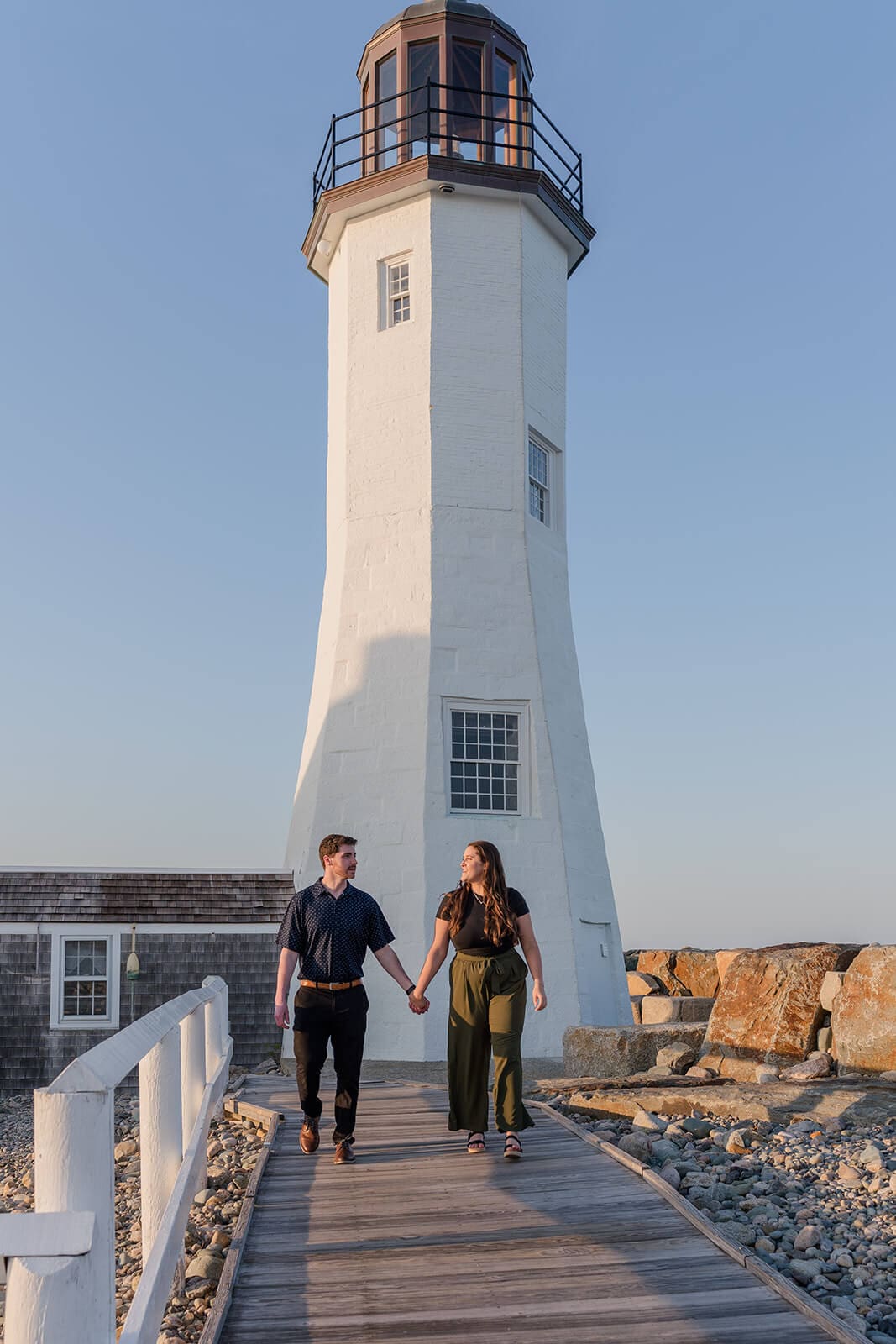 Engaged couple walking hand in hand along the boardwalk beside Scituate Lighthouse