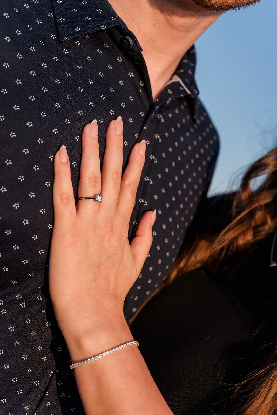 Close-up of a woman’s diamond engagement ring as her hand rests against her fiancé’s shirt