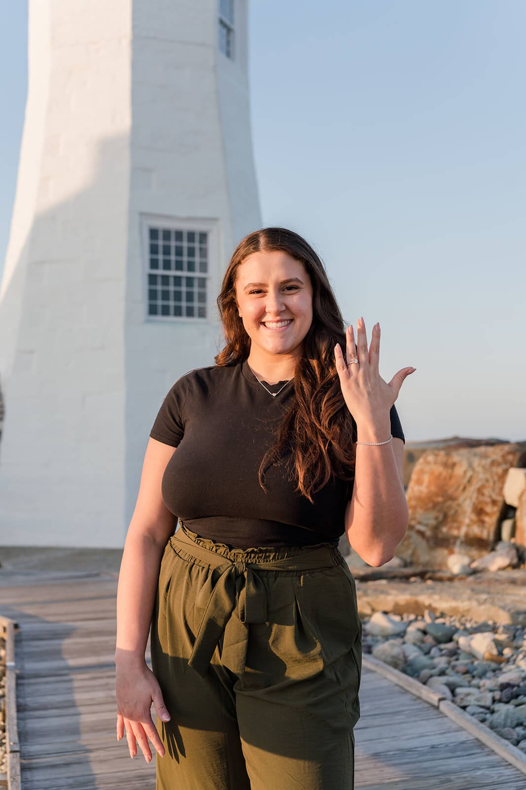 Woman smiling and holding up her engagement ring with Scituate Lighthouse behind her