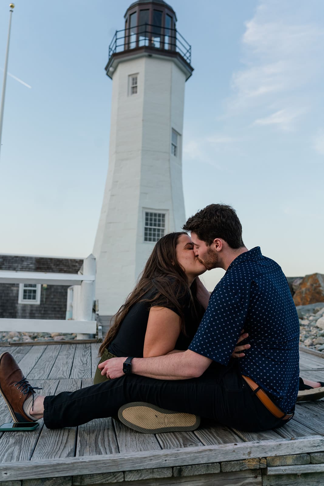 Couple sitting together and kissing on a wooden platform near Scituate Lighthouse