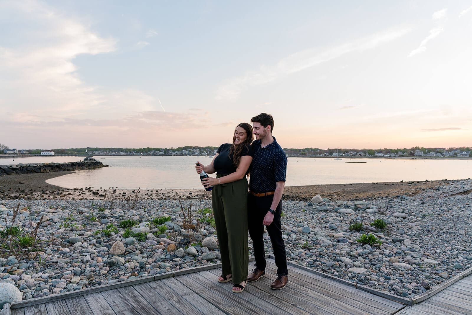 Newly engaged couple standing together on a waterfront boardwalk at sunset popping a bottle of champagne