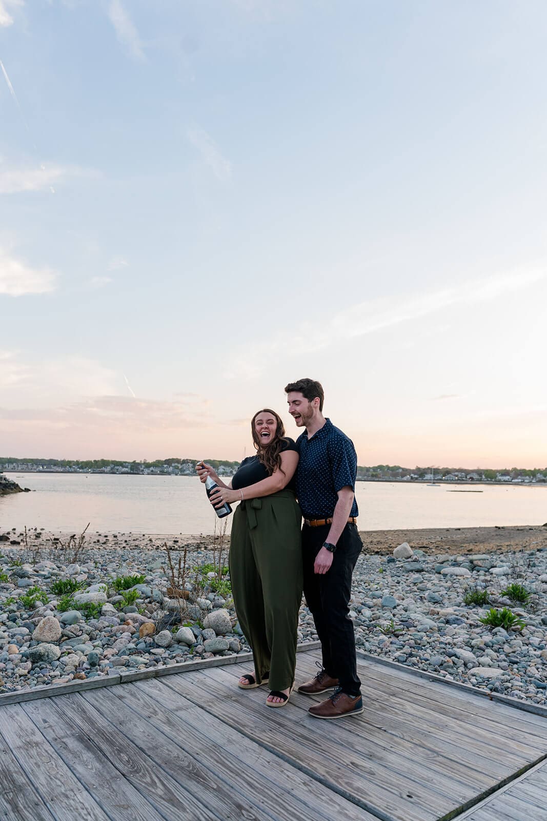 Couple standing together on a boardwalk overlooking the rocky shoreline and water at sunset popping a bottle of champagne