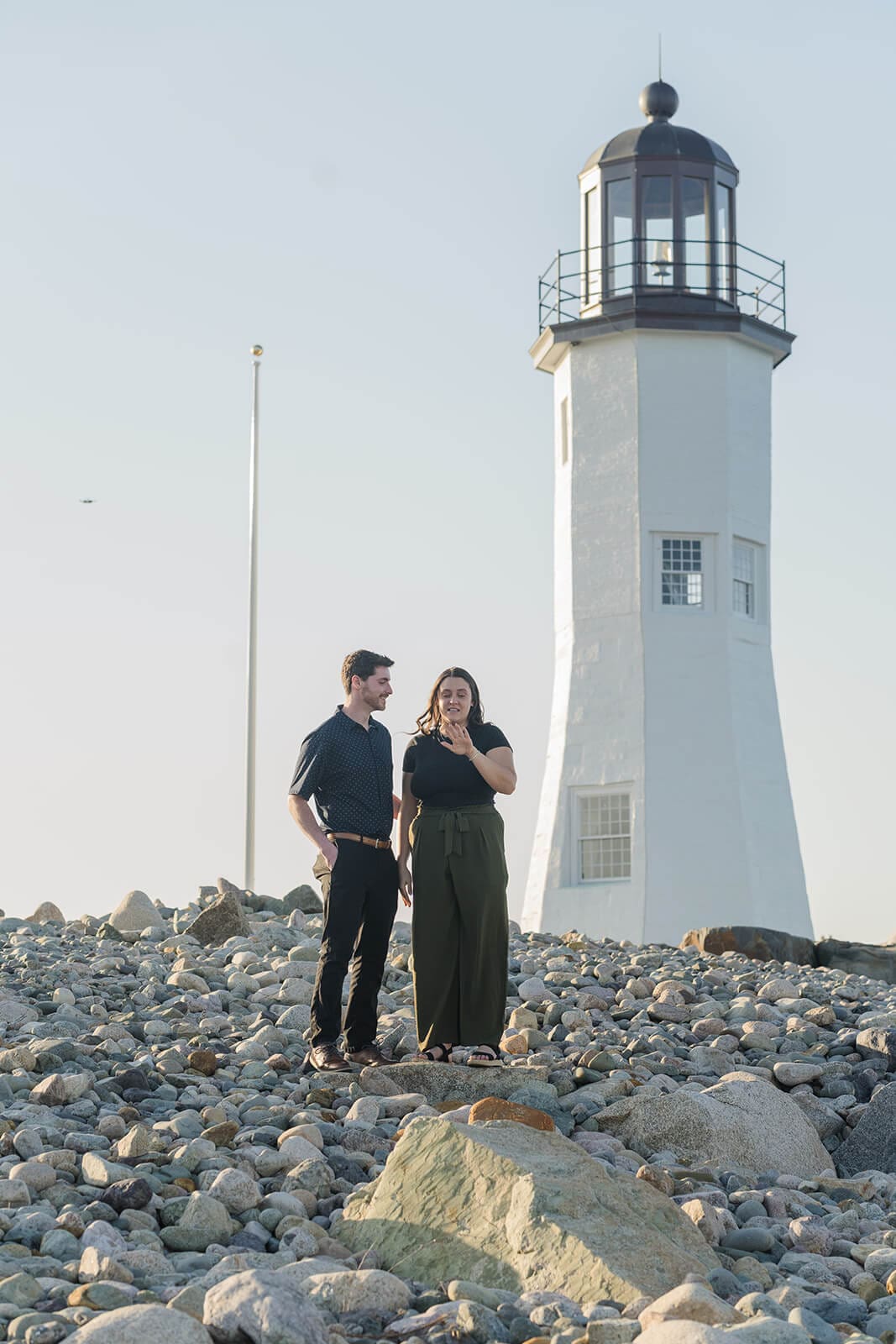 Engaged couple smiling at each other among the rocks beneath Scituate Lighthouse