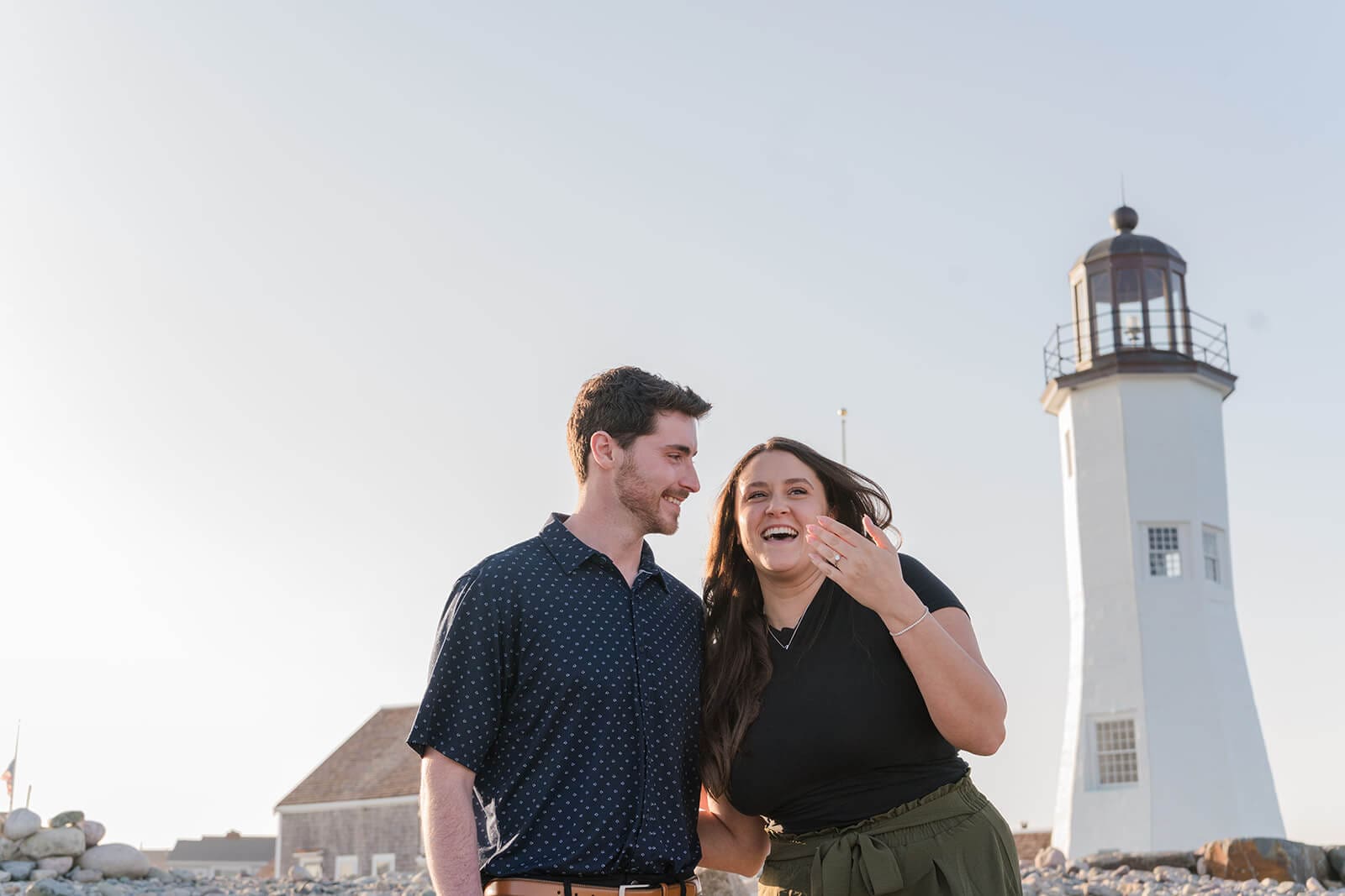 Woman laughing and showing her engagement ring beside her fiancé at Scituate Lighthouse