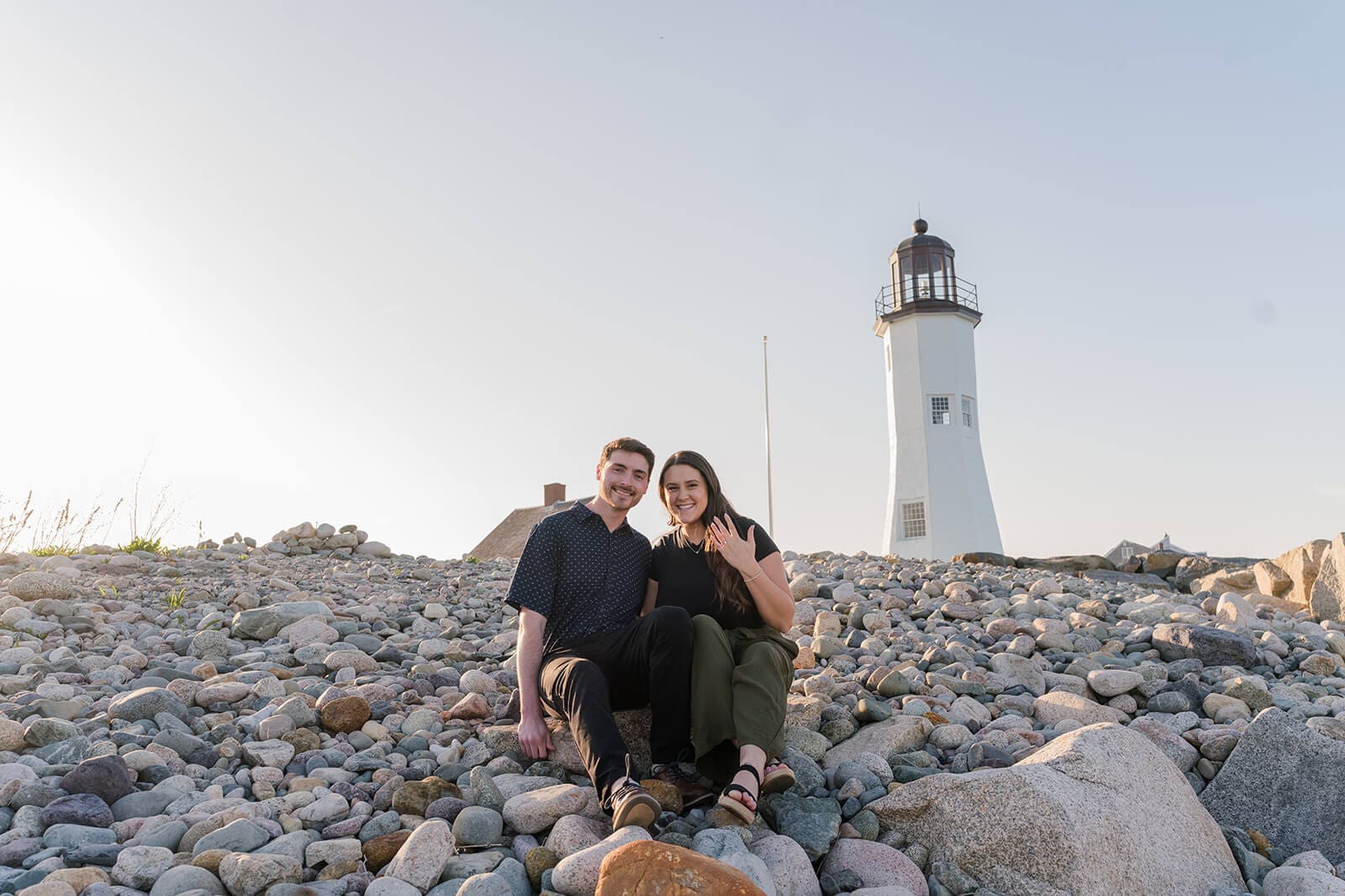 Engaged couple sitting together on the rocks with Scituate Lighthouse in the distance