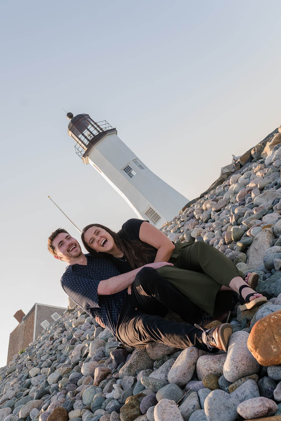 Couple sitting together on the rocks and smiling with Scituate Lighthouse behind them